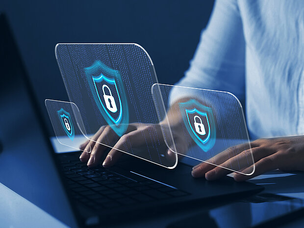 Eine Hand tippt auf einen Computer, während drei transparenten Schlössern, welche für die Cyber Security stehen, über dem Computer schweben